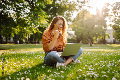 Beautiful woman in casual clothes on a green lawn with a laptop. Happy freelancer woman working on laptop outdoors. Education and freelancing concept.