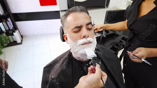 Client in hairdressing shop making faces during beard dyeing process with two stylists.MOV photo