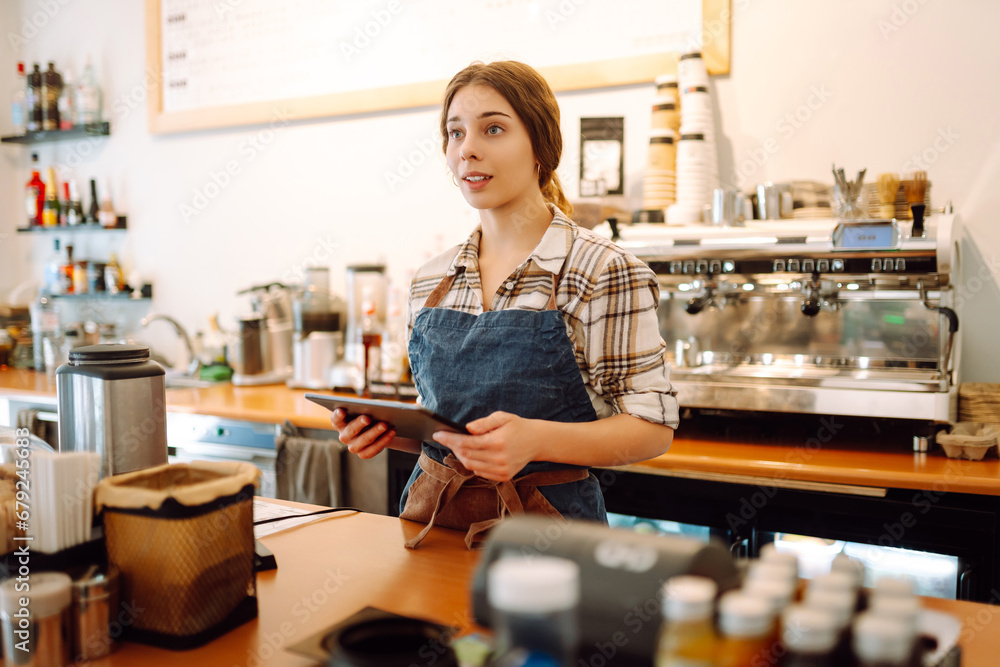 Portrait of a friendly female barista holding a tablet computer and smiling. A young coffee shop owner standing behind the bar using a digital tablet. Takeaway concept, technology. Portrait of a friendly female barista holding a tablet computer and smiling. A young coffee shop owner standing behind the bar using a digital tablet. Takeaway concept, technology.