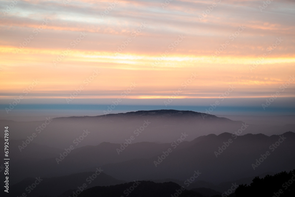 Golden sunset illuminates mountains, clouds, and a tranquil forest, casting a warm orange embrace over the serene landscape.