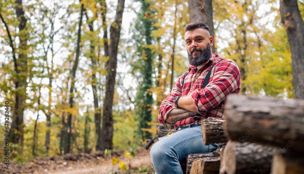A hipster lumberjack relaxes on the pile of freshly cut logs, pausing for a portrait.