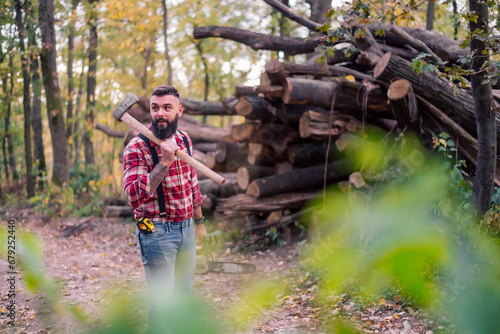 Portrait of a stylish hipster confidently navigating the forest path with an axe and chainsaw in hand, headed towards a stack of cut logs.
