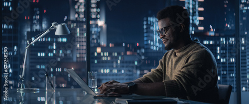 Successful Handsome Black Businessman Working on Laptop Computer in Big City Office in the Evening. Finance Investment Manager Checking Financial Data from Project Management Report.