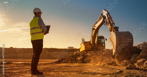 Caucasian Male Civil Engineer Using Laptop Computer On Construction Site Of New Real Estate Project. Man Observing Industrial Heavy Machinery At Work, Laying Foundation For Apartment Building. Sunny photo