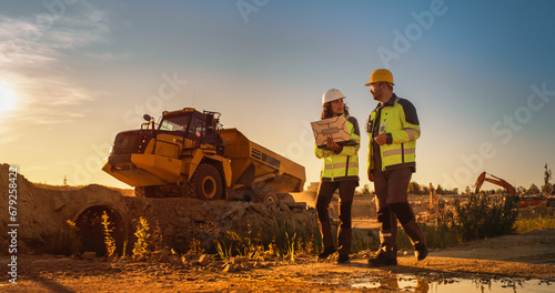 Caucasian Male Architect And Hispanic Female Urban Planner Walking On Construction Site With Laptop Computer And Talking About New Real Estate Project. Construction Truck Driving By. Golden Hour