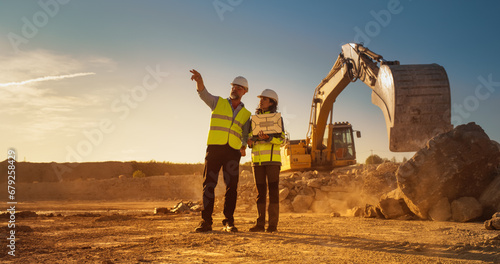 Caucasian Male Civil Engineer And Hispanic Female Architect Walking On Construction Site With Laptop Computer And Talking About New Real Estate Project. Machinery Working On Background. Golden Hour. photo
