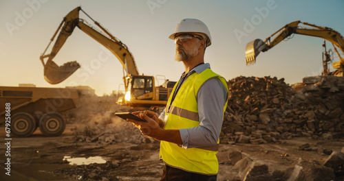 Caucasian Male Civil Engineer Wearing Protective Goggles And Using Tablet On Construction Site On Sunny Day. Man Inspecting Building Progress. Excavator Loading Materials Into Big Industrial Truck. photo