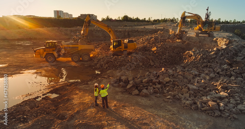 Aerial Drone Shot Of Construction Site On Sunny Evening: Industrial Excavators Digging Rocks And Loading Them Into A Truck. Engineer And Architect Observing Process, Discussing Real Estate Project