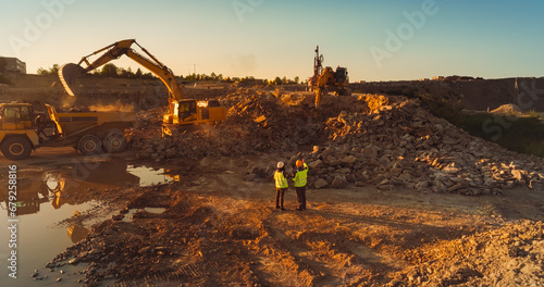 Aerial Drone Shot Of Construction Site On Sunny Day  Industrial Excavators Digging Rocks And Loading Into A Truck. Engineer And Inspector Observing Process  Discussing New Real Estate Project