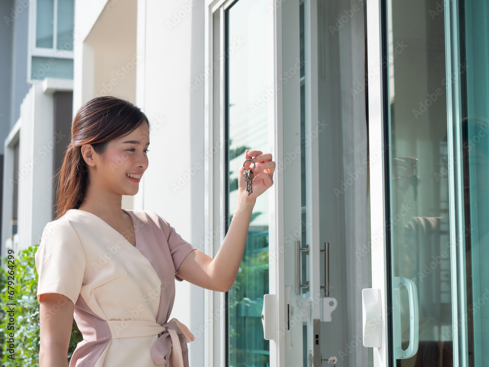 Happy Asian woman smiling and holding keys in front of the door of a new house.