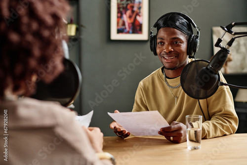 Young African American man with paper speaking in microphone while explaining something or asking question to guest invited for interview