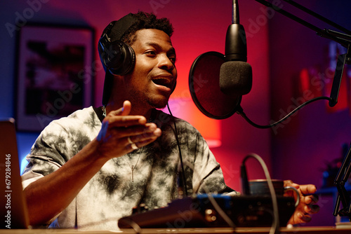 Young male radio presenter in headphones talking in microphone while communicating with invited guest in studio illuminated by neon light