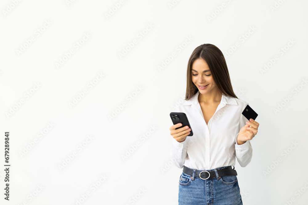 Young woman holding credit card and looking at smartphone app in phone application, standing over white background Young woman holding credit card and looking at smartphone app in phone application, standing over white background