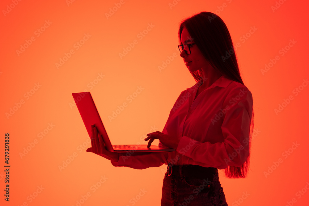 Portrait of young woman posing, having video conference on laptop isolated over red background in neon light.