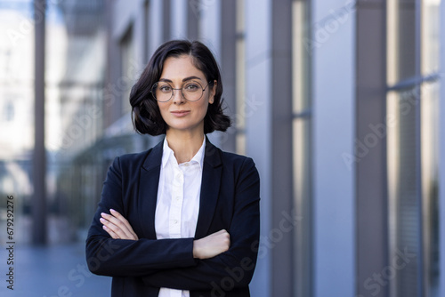 Portrait of a smiling young female lawyer standing outside the courthouse in a business suit, arms folded across her chest, smiling and confidently looking at the camera