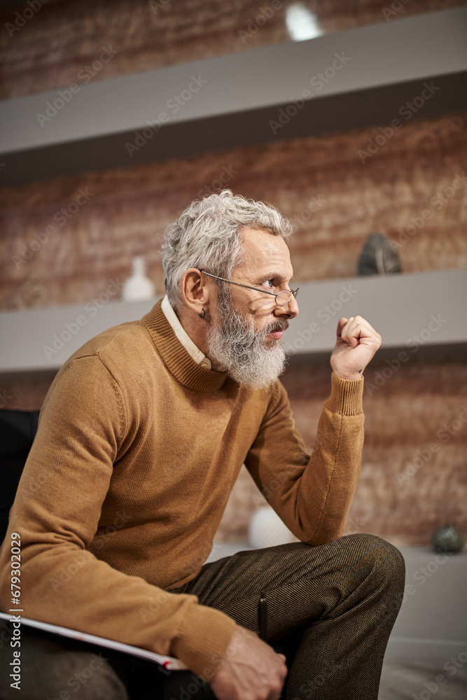 bearded middle aged psychologist in glasses holding pen near notebook and listening during session bearded middle aged psychologist in glasses holding pen near notebook and listening during session