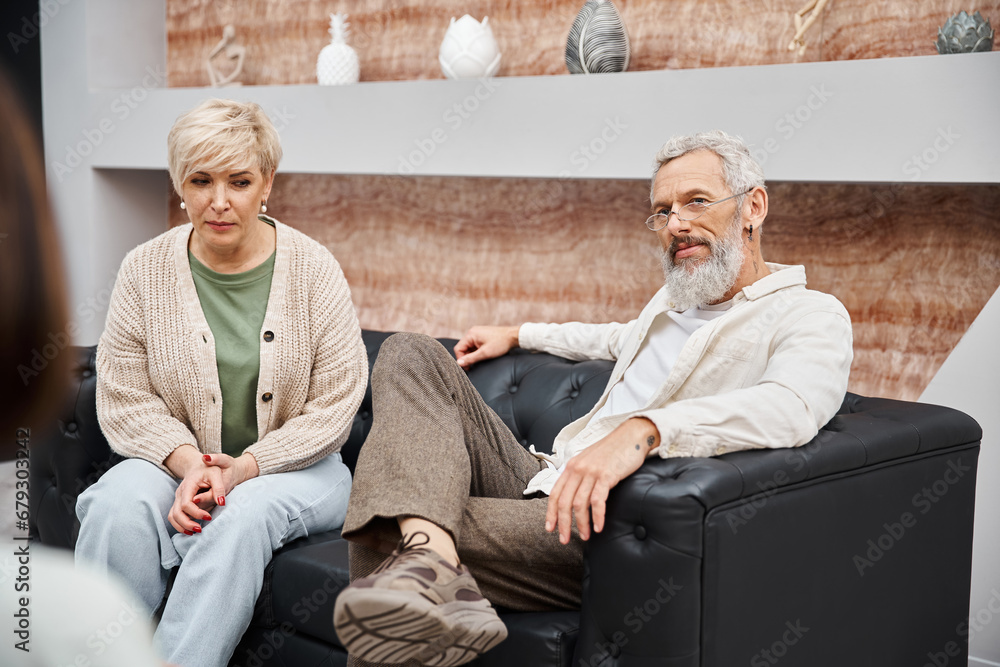middle aged couple sitting on leather couch and looking at family counselor during consultation middle aged couple sitting on leather couch and looking at family counselor during consultation
