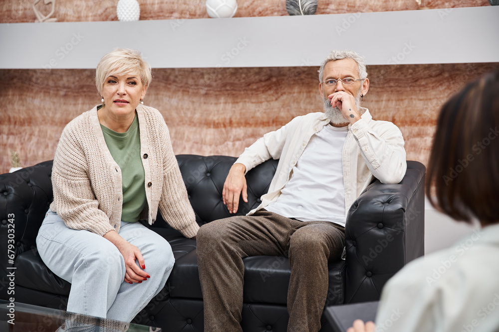 middle aged couple sitting on leather couch and looking at psychologist during consultation middle aged couple sitting on leather couch and looking at psychologist during consultation