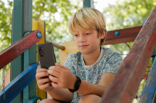 10 years old cute blonde boy using smartphone outdoors. Portrait of concentrated kid sitting in a playground alone holding mobile phone looking social media. Rest, education and technology concept. photo