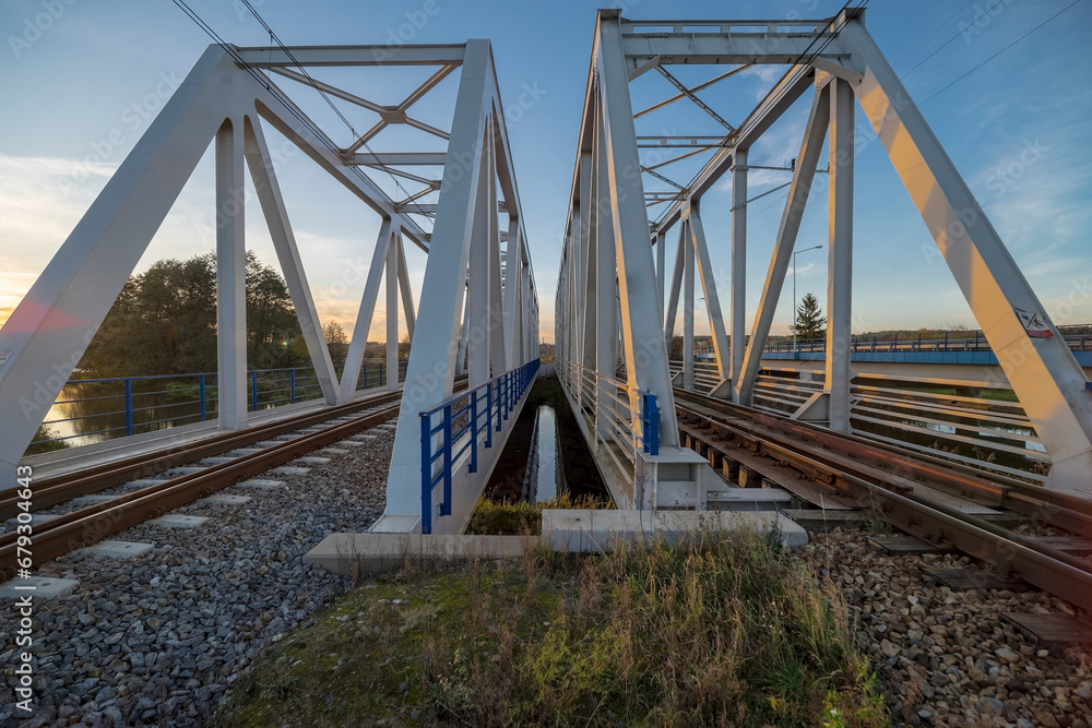 Dwudzielny stalowy most kolejowy o ażurowej konstrukcji w porze zachodu słońca w listopadowe popołudnie nad rzeką Kamienną .