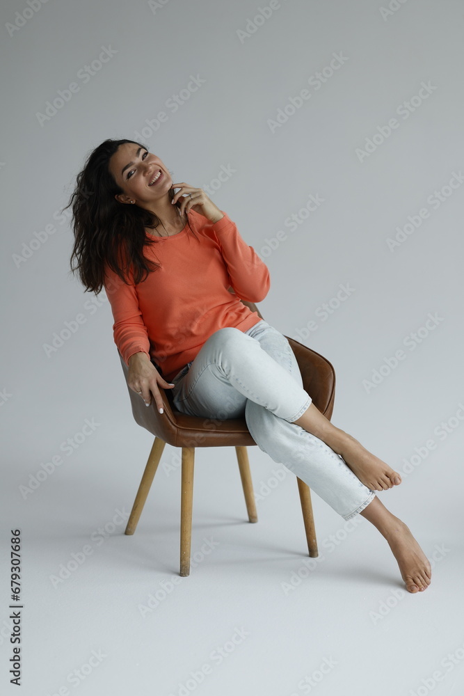 Beautiful woman of 30 years old sitting on chair, white background. Portrait of brunette girl smile. Orange sweater and light jeans. Woman barefeet, thoughtful. Beautiful woman of 30 years old sitting on chair, white background. Portrait of brunette girl smile. Orange sweater and light jeans. Woman barefeet, thoughtful.