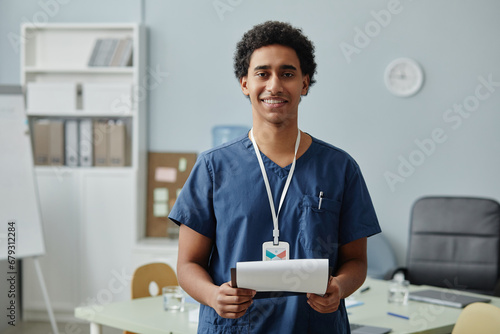 Waist up portrait of young Arabian man wearing scrubs uniform and holding clipboard in clinic, copy space photo