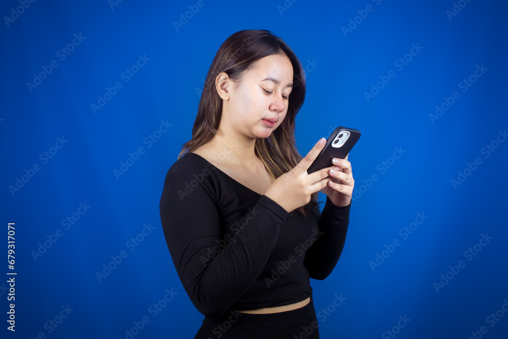 A young Asian woman is happy and smiling while using a smartphone on a blue background A young Asian woman is happy and smiling while using a smartphone on a blue background