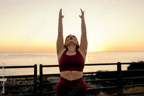 Plus-size woman in a yoga pose feeling empowered against a beautiful sunset backdrop, promoting a body-positive lifestyle. photo