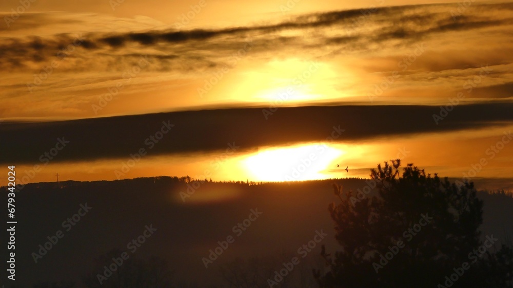 Orange morning sky with clouds, yellow sunlight during sunrise over dark horizon of Silesian Beskid mountains in Poland with forests, Christian cross and birds in flight.