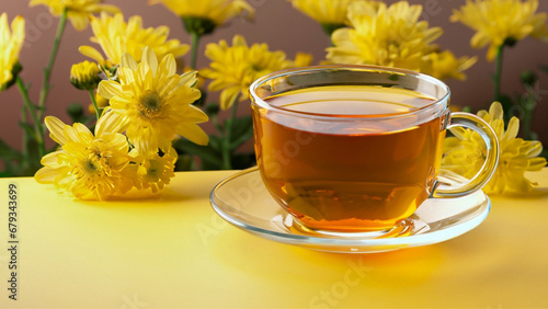 Uma xícara de vidro transparente com chá, sobre uma superfície amarela e muitas flores amarelas ao fundo.