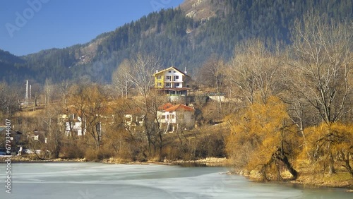 Zoom out Frozen lakeside and beautiful mountain village houses nestled among colorful trees displaying foliage. 4K Landscape at Plovdiv Rhodope Mountains, Smolyan in Bulgaria
 photo