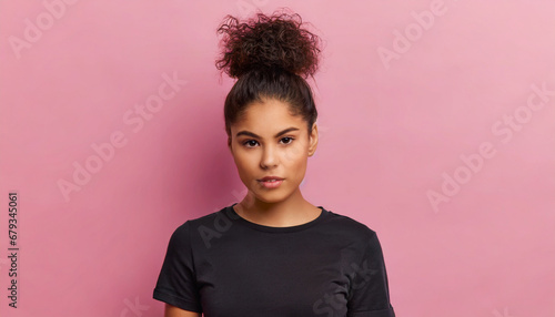 serious displeased latin woman with hair bun raises eyebrows looks attentively at camera purses lips has dimple on cheek dressed in casual black t shirt and jeans isolated over pink background photo