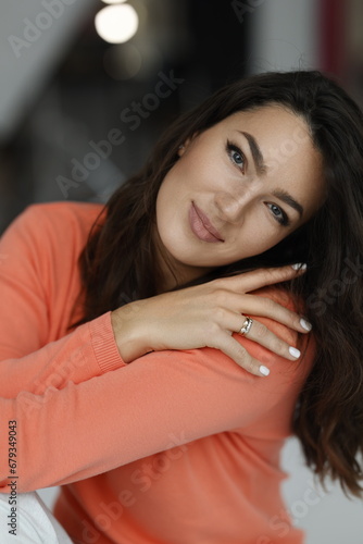 Beautiful woman of 30 years old sitting on chair, white background. Portrait of brunette girl smile. Orange sweater and light jeans. Woman barefeet, thoughtful.