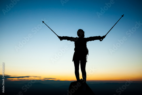 Silhouette of a hiker girl on a rock pedestal with hands up. Beautiful orange sunset on blue sky.