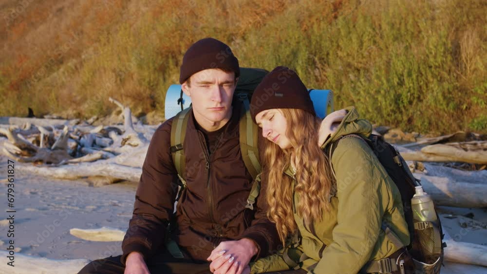 Young loving couple with backpacks sits on log on river beach in autumn. Hikers rest on lake bank. Boyfriend and girlfriend doing hiking in nature. Tourists relaxing during sunset Young loving couple with backpacks sits on log on river beach in autumn. Hikers rest on lake bank. Boyfriend and girlfriend doing hiking in nature. Tourists relaxing during sunset