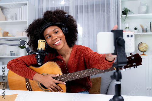 Host channel of beautiful African woman influencer setting smartphone, sing with play guitar in broadcast. Time slot of music blogger on live social media online. Concept of audio creator. Tastemaker. photo