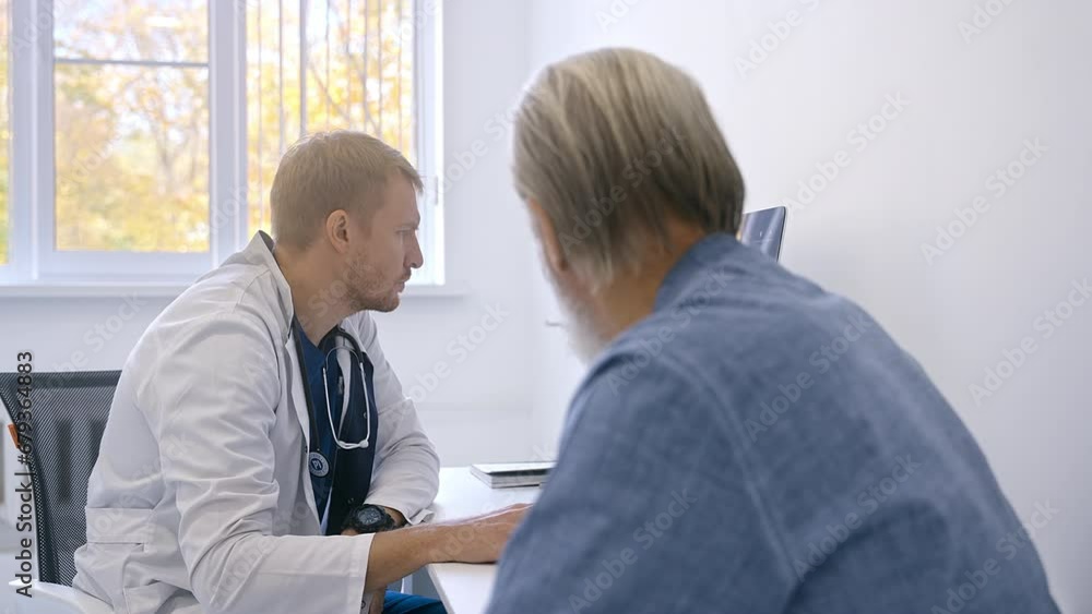 Doctor in blue clothes and white coat measuring heartbeat of stylish elderly patient with beard in office of modern medical facility Doctor in blue clothes and white coat measuring heartbeat of stylish elderly patient with beard in office of modern medical facility