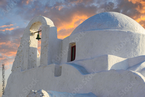 Sunset starts on the island of Mykonos with a white-washed church with a lone bell aglow in the foreground. 