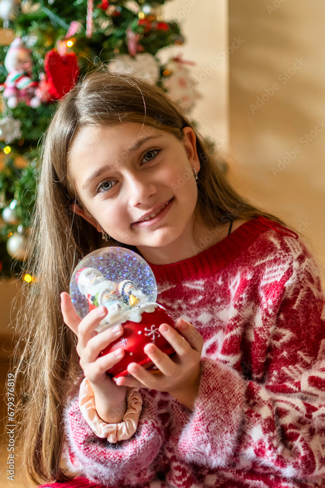 Naklejka premium Radiant 8-Year-Old Girl, Joyful with a Snow Globe by the Christmas Tree at Home