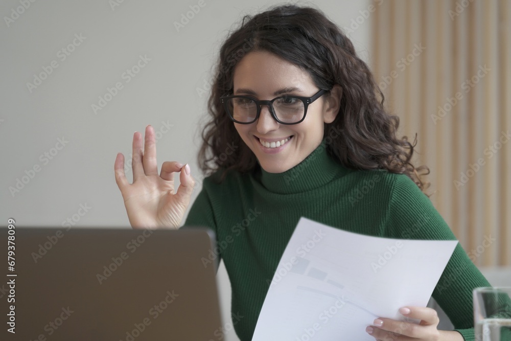 Confident woman in glasses giving OK sign during a video call, with papers and laptop Confident woman in glasses giving OK sign during a video call, with papers and laptop