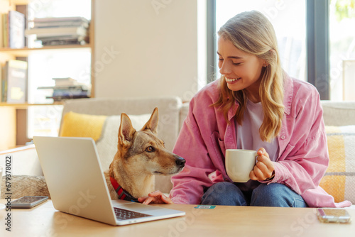 Smiling beautiful woman using laptop holding cup of coffee working in modern cafe with adorable cute dog. Successful happy freelancer sitting at workplace near lovely pet 