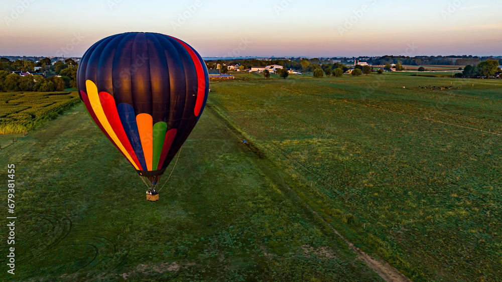 Obraz premium An Aerial View of a Black Striped Hot Air Balloon Floating Low Over Fields and Farmlands in Rural America on a Sunny Summer Morning.