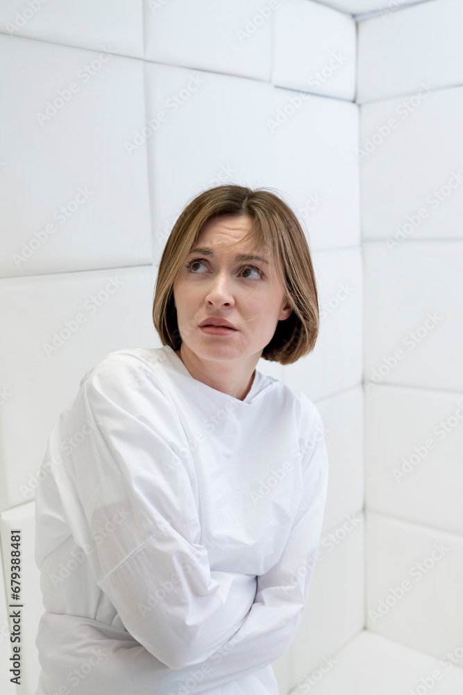 Young frustrated girl in a straitjacket is in an isolated white room in a mental hospital