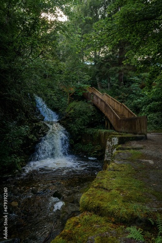 a bridge spanning over a small waterfall in a wooded area © Wirestock