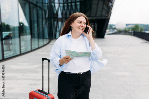Beautiful redhead woman holding paper map and talking on smartphone outdoors. Lady with suitcase choosing route via paper map, navigating her urban journey © mdyn