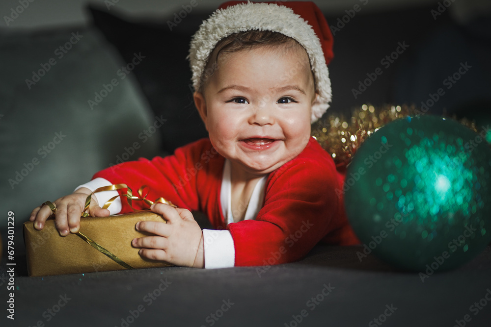 Obraz premium happy smiling baby boy in red santa claus costume lying with christmas hat and gifts or presents at home in the living room