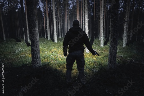 Silhouette of a man with a pistol and a flashlight in a dark forest, viewed from the back. photo