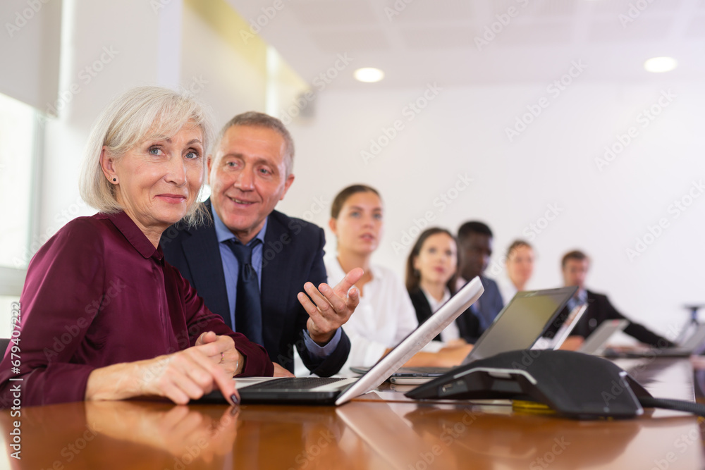 Fototapeta premium Positive senior woman manager sitting at desk beside colleagues and using laptop during conference.