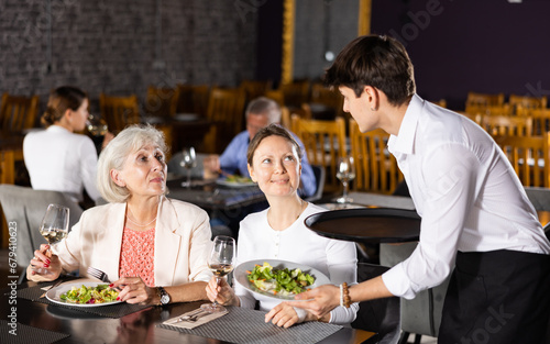 Young male waiter brought food to order of young girl and elderly mother to visitors of restaurant. High quality photo
