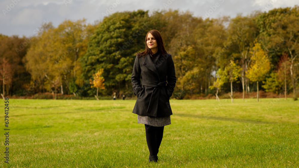 Full length shot of a middle-aged woman with medium length brown hair, walking in a park in the autumn with trees behind. Full length shot of a middle-aged woman with medium length brown hair, walking in a park in the autumn with trees behind.